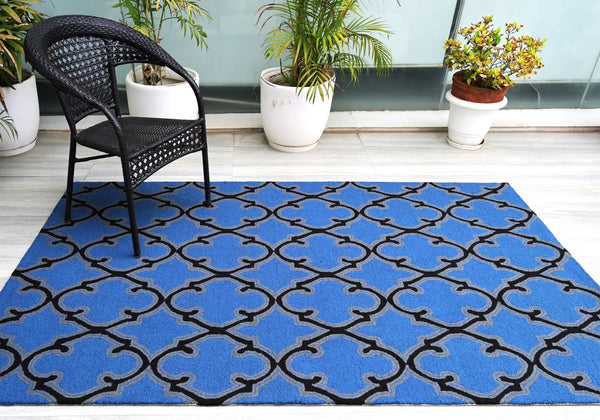 Bluenox Rug styled in a modern open space with a wooden chair and green plants, showcasing its deep blue tone and black geometric pattern in natural daylight.