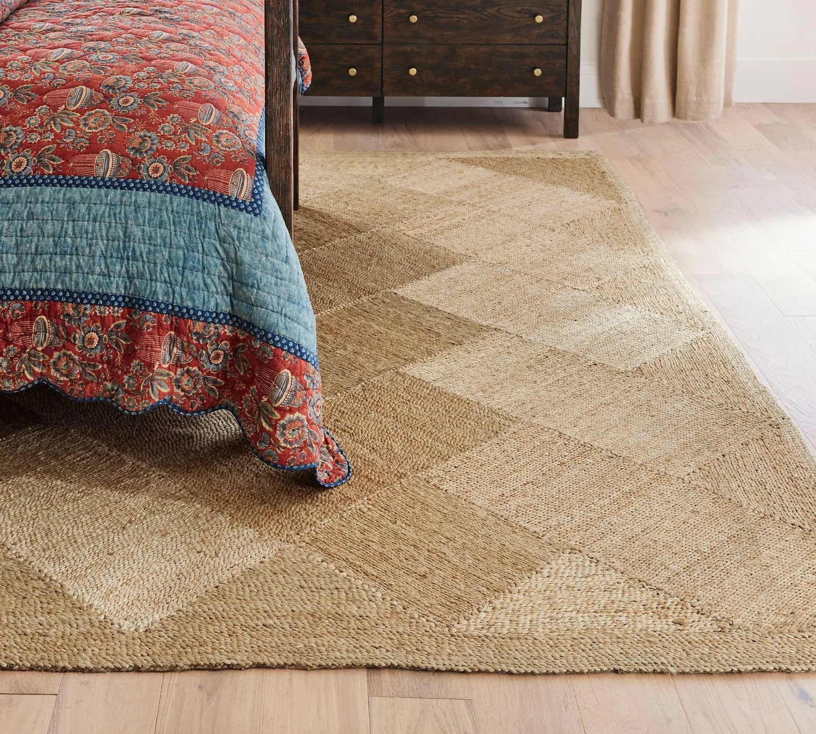 Cascara Rug placed beside a patterned bedspread in a cozy bedroom, showcasing its geometric jute weave and natural earthy tones on wooden flooring.