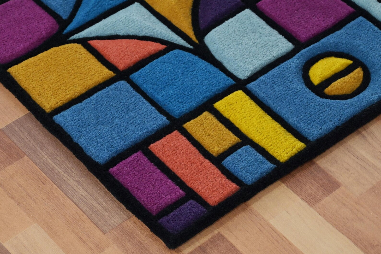Corner detail view of Chromatic Chaos Rug showing colorful geometric blocks in blue, yellow, and pink with bold black outlines on wooden flooring