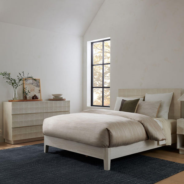 Driftstone Rug placed under a beige bed in a minimal bedroom setting, enhancing the warm and cozy ambiance with its deep textured weave.
