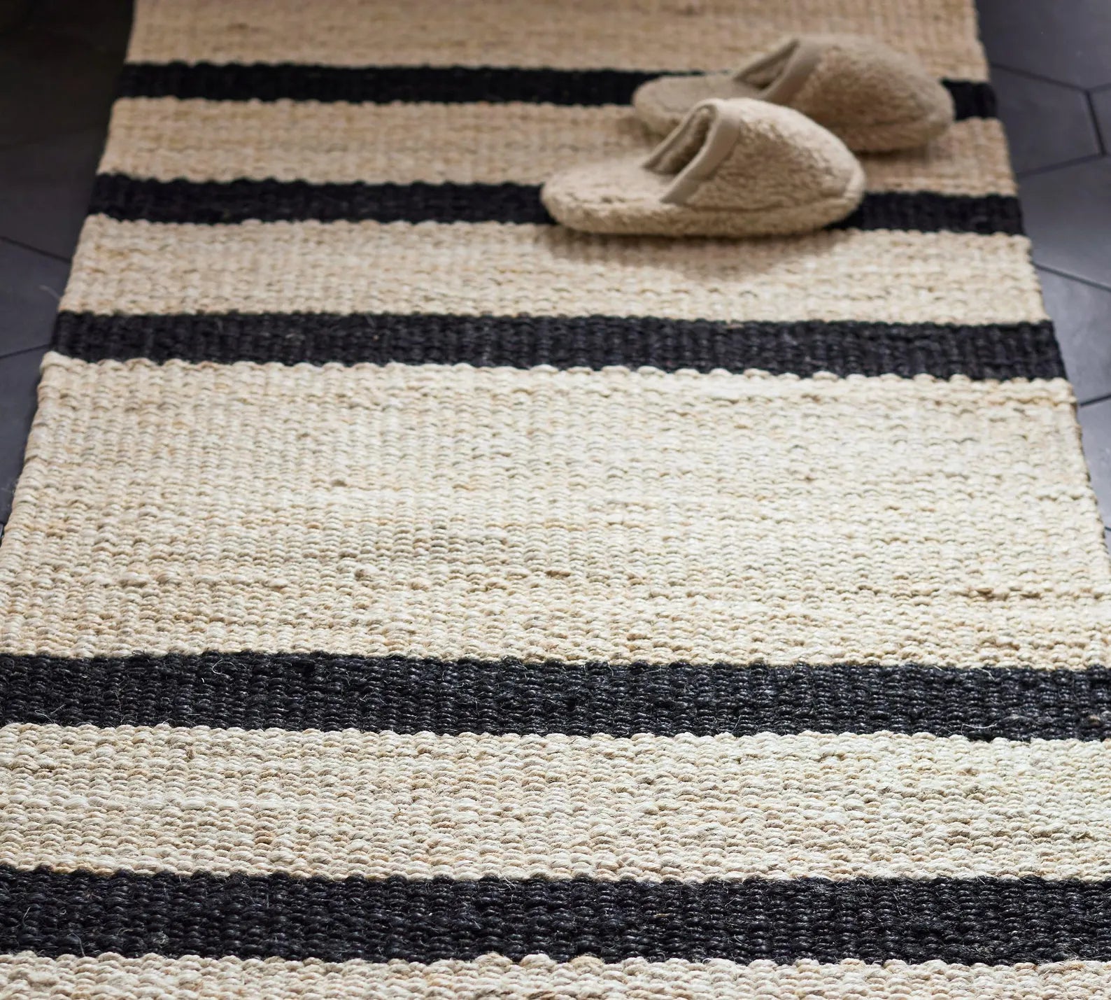 Noirpath Rug styled on a tiled floor with black and beige handwoven stripes, featuring slippers for a cozy lifestyle scene.