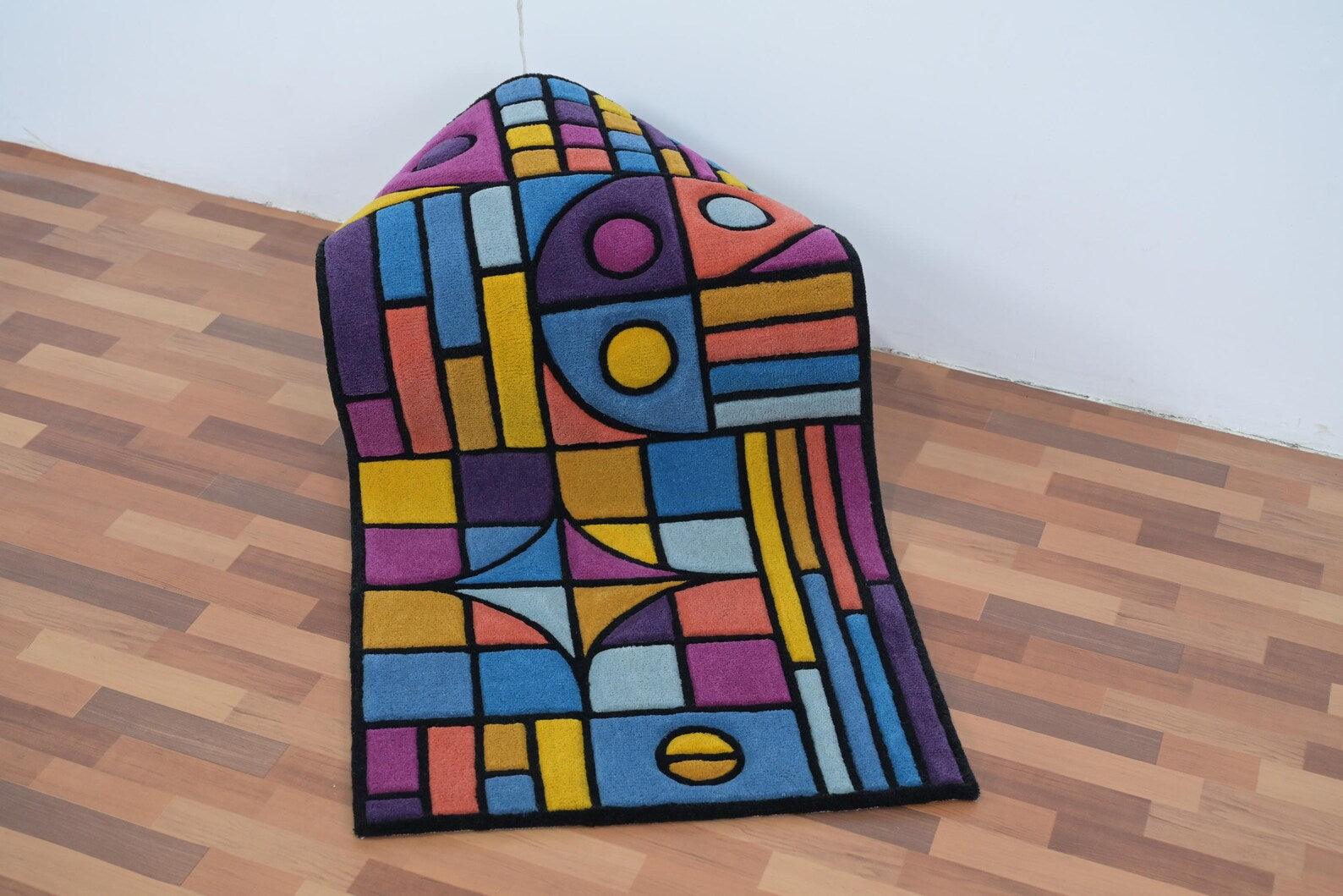 Rolled display view of Chromatic Chaos Rug showing vibrant abstract geometric pattern in blue, yellow, pink, and orange tones on wooden floor background