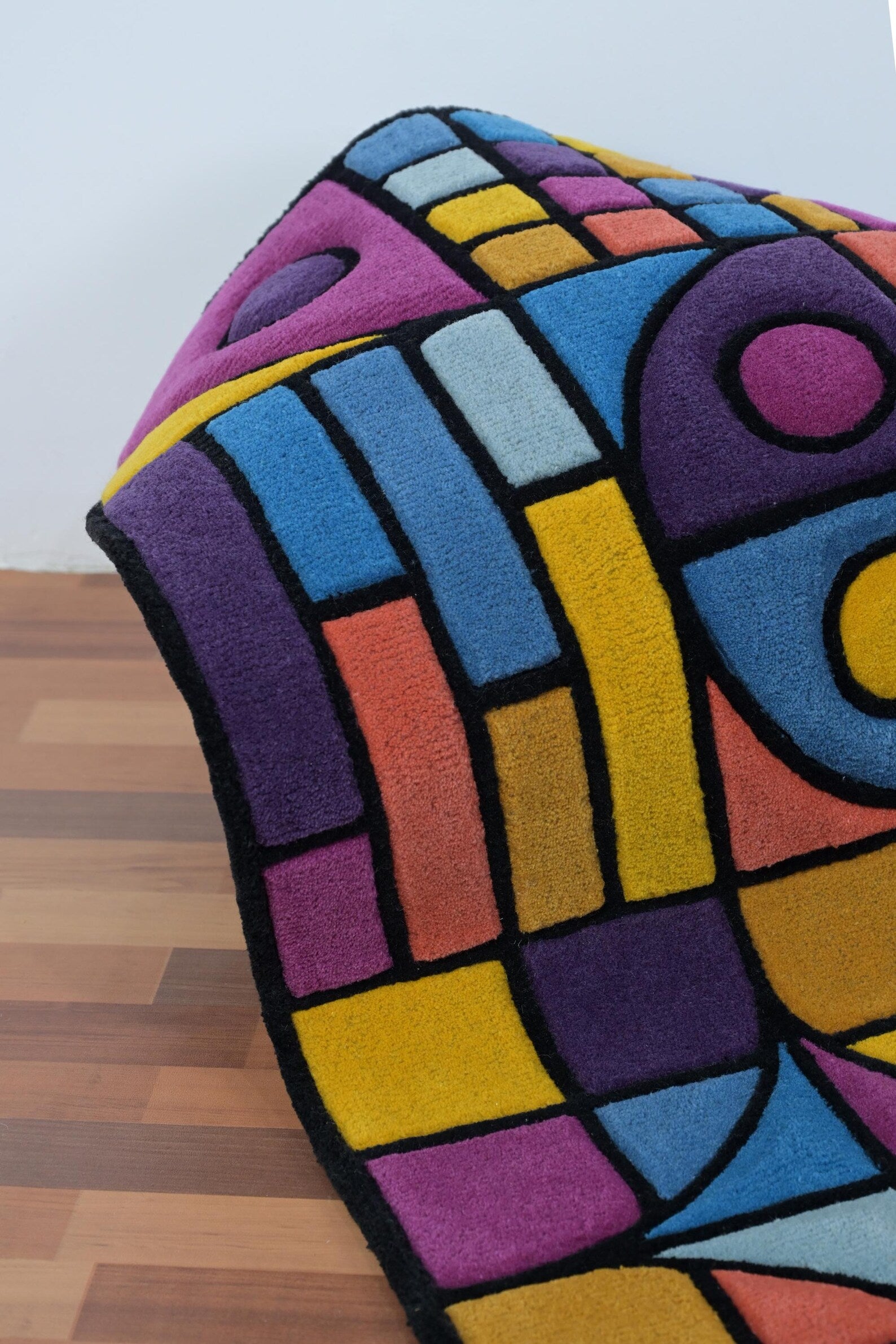 Rolled edge view of Chromatic Chaos Rug showing bright geometric blocks in blue, purple, yellow, and orange with black outlines on wooden flooring