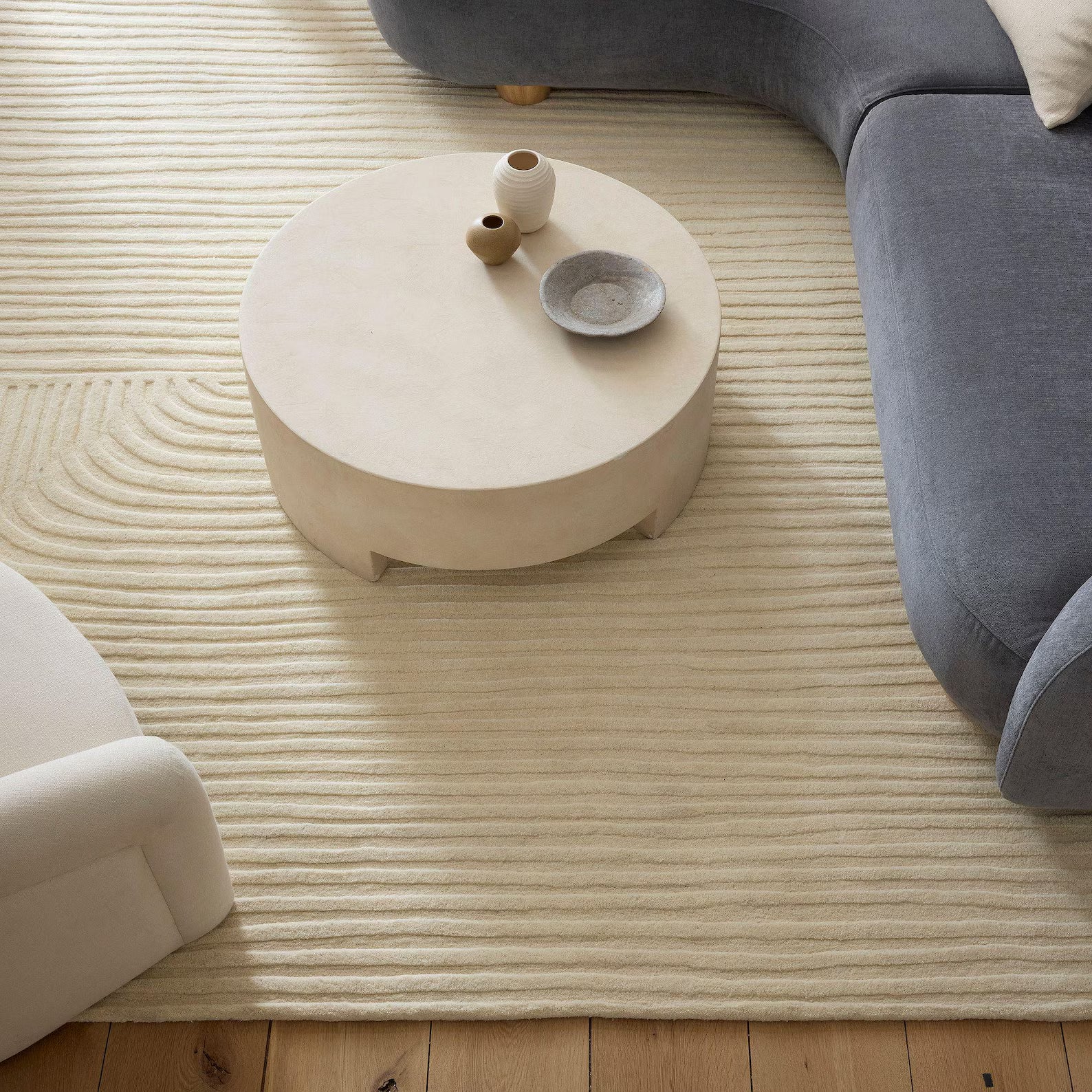 Minimal living room with Softwave Rug placed under a round beige coffee table beside a soft grey sofa, showing gentle wave-like tufted texture.