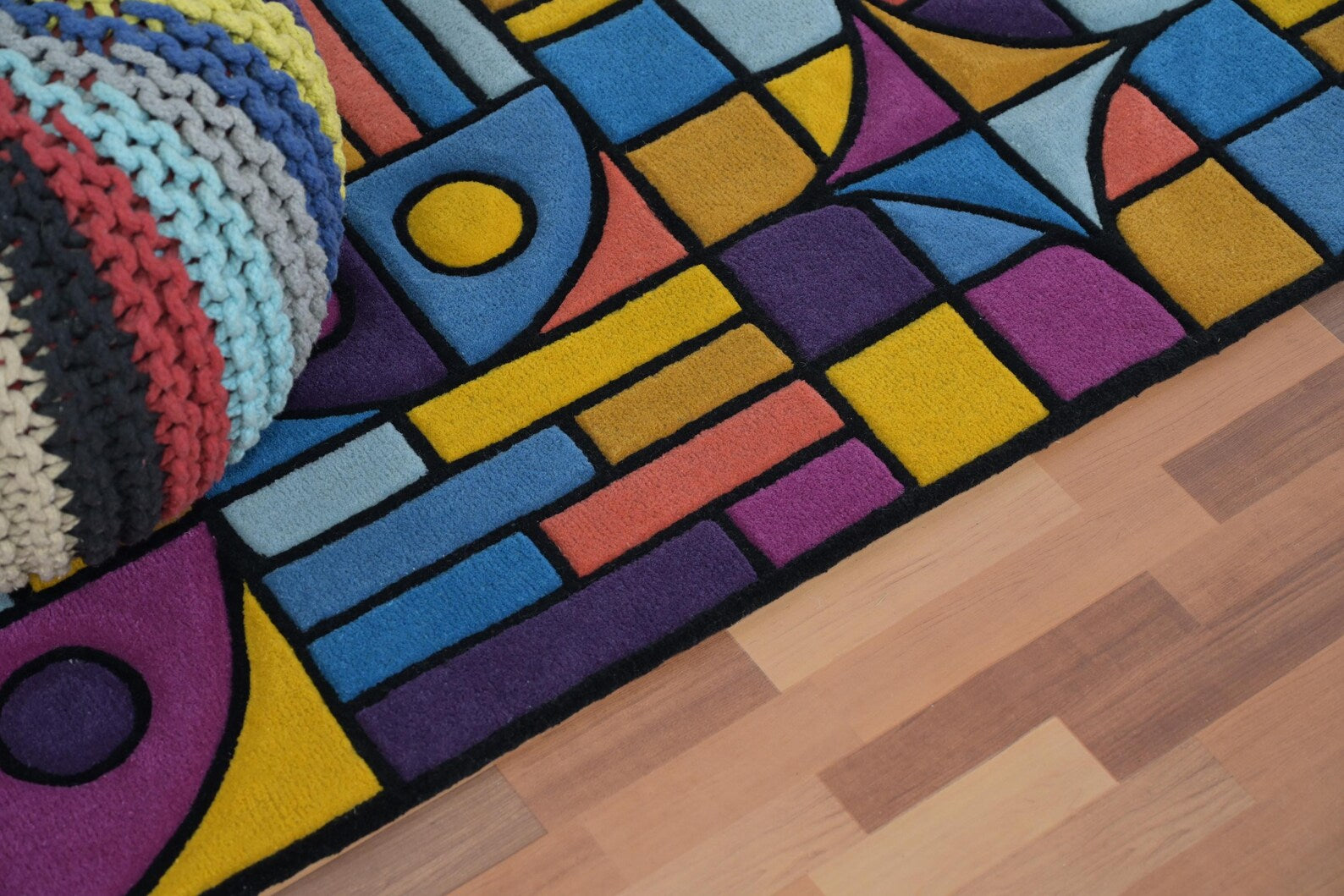 Top angle detail view of Chromatic Chaos Rug showing abstract geometric shapes in yellow, blue, purple, and orange tones with black outlines on wooden flooring