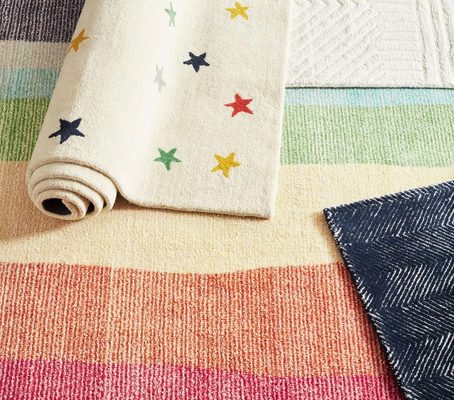 Folded view of Twinkle Rug showing colorful star motifs on ivory wool, placed alongside striped rugs in soft pastel tones highlighting its playful yet elegant contrast.