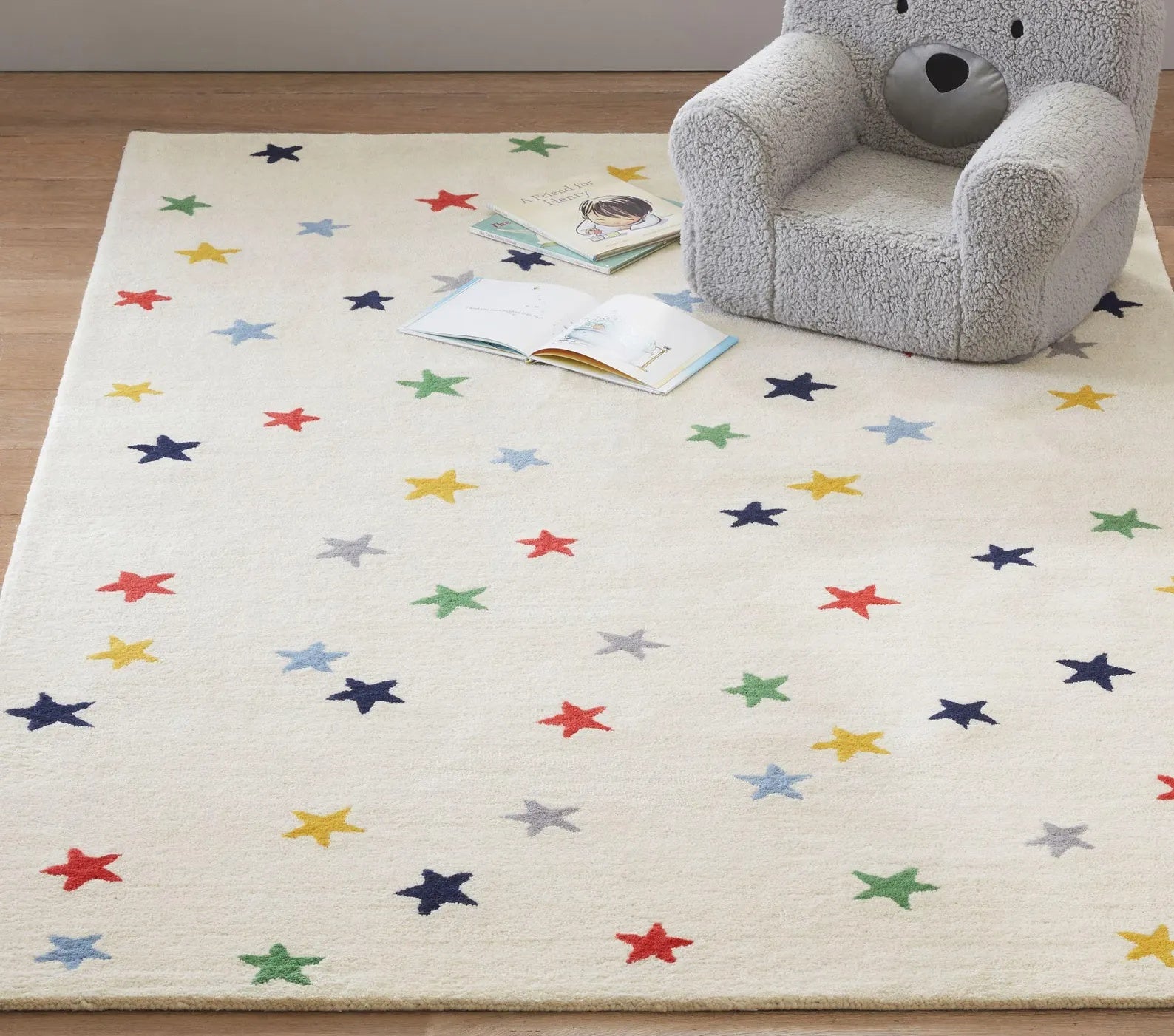 Twinkle Rug placed in a cozy nursery with a grey bear chair and open books on top, featuring colorful stars on a soft ivory wool base adding warmth and playful charm to kids’ spaces.