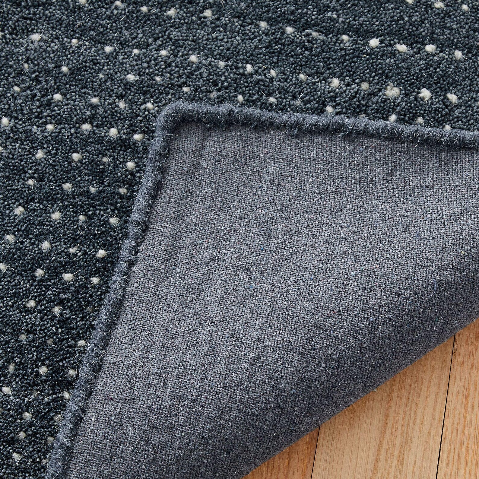 Angled close-up of folded hand tufted Indigo Dot Texture Area Rug showing its fine woven pattern and smooth edges on a light wooden floor.