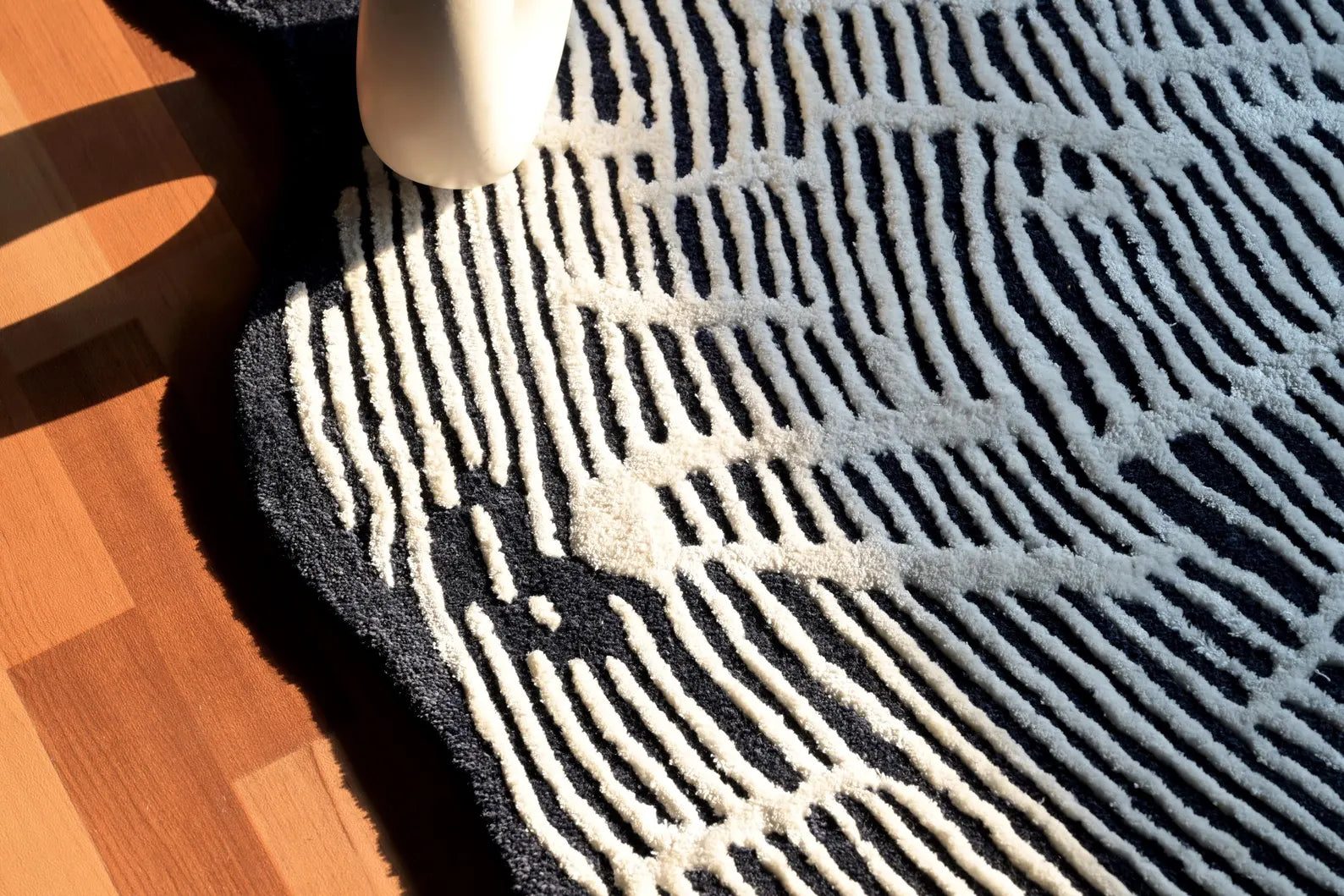 Hand tufted Sequoia Rug corner view under natural sunlight highlighting its white tree-ring pattern, dark wool texture, and curved organic edge on wooden flooring.
