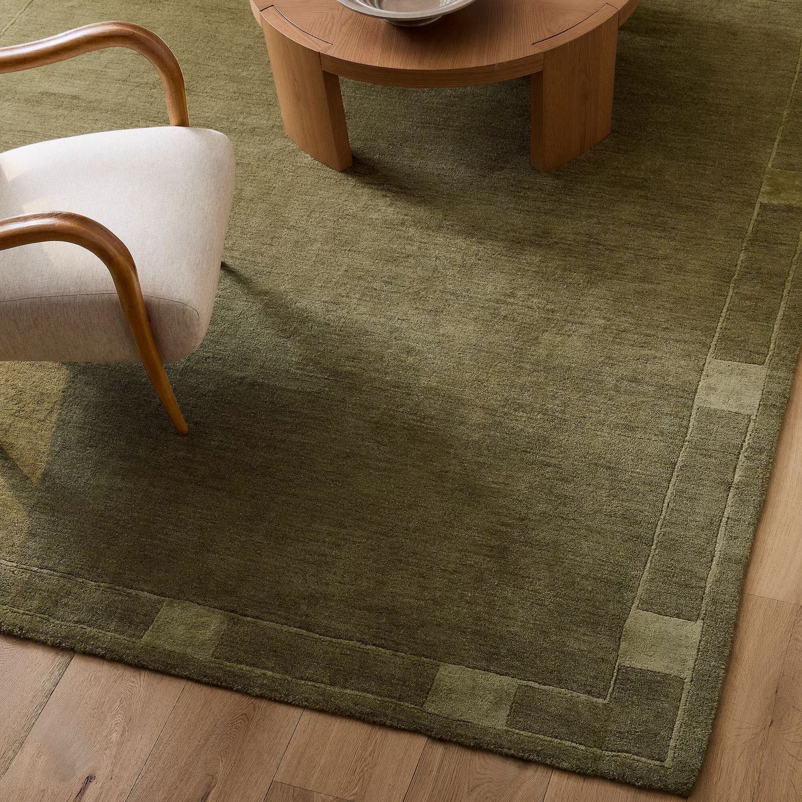 An olive mid-century modern hand tufted rug placed beneath a curved chair and round wooden table in a beige light-brown interior.